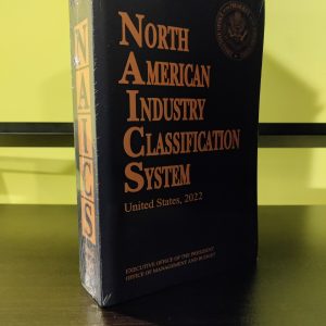 A sealed hardcover book titled "U.S. NAICS Manual - Hardcover 2022-2027" rests on a dark surface against a yellow background.