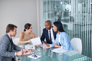 Four business professionals sit at a glass table in an office, discussing documents and taking notes during a meeting.