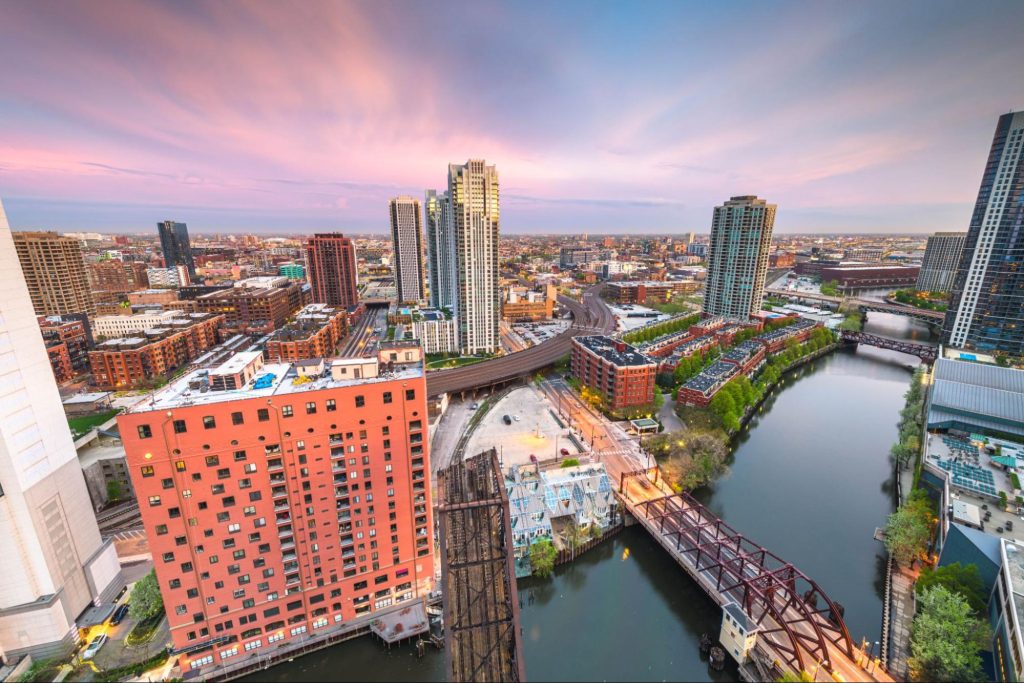 Aerial view of a cityscape at sunset with high-rise buildings, a river, a bridge, and a highway curving through the scene.
