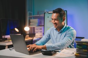 A man in a blue shirt sits at a desk, smiling while working on a laptop, possibly checking his company's DUNS number in an office with shelves and documents in the background.